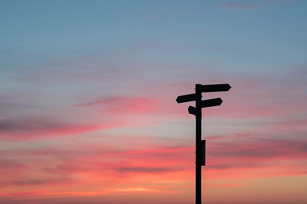 A black pole with arrows in different directions against a blue, yellow and pink sky.