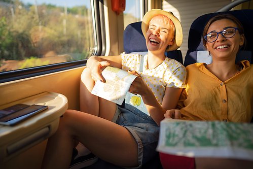 Two people sitting on a train. They laugh and each have a map in their hands.