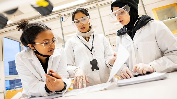 Tre kvinnliga studenter med skyddsglasögon diskuterar i ett labb.